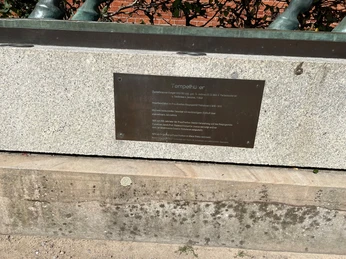 Schwarze Gedenktafel auf einer Steinmauer mit Inschrift und Metallgeländer darüber. Klarer Himmel.