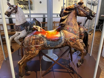 Drei kunstvoll geschnitzte, verzierte hölzerne Karussellpferde stehen in einem musealen Ambiente.Three ornately carved, decorated wooden carousel horses stand in a museum-like setting.Tre kunstfærdigt udskårne, dekorerede karruselheste af træ står i museumslignende omgivelser.Drie sierlijk gesneden, versierde houten carrouselpaarden staan in een museumachtige omgeving.