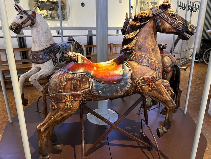 Deutsches Pferdemuseum Verden Drei kunstvoll geschnitzte, verzierte hölzerne Karussellpferde stehen in einem musealen Ambiente.Three ornately carved, decorated wooden carousel horses stand in a museum-like setting.Tre kunstfærdigt udskårne, dekorerede karruselheste af træ står i museumslignende omgivelser.Drie sierlijk gesneden, versierde houten carrouselpaarden staan in een museumachtige omgeving.