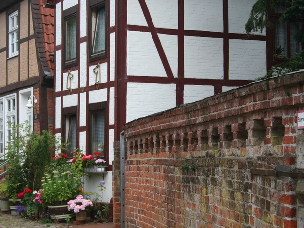 Fachwerkhäuser und historische Backsteinmauer in einer Straße, gesäumt von Blumentöpfen.