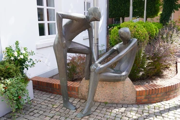 Skulptur vor VGH Moderne Bronzeplastik zweier stilisierter Figuren auf einem Granitstein, umrahmt von Pflanzen und Büschen.