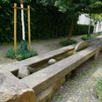Langer, steinerner Brunnen mit flachem Wasserbett verläuft durch eine gepflasterte Gartenlandschaft.