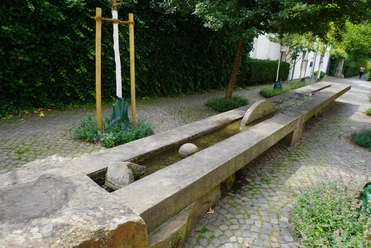 Brunnen Verden Langer, steinerner Brunnen mit flachem Wasserbett verläuft durch eine gepflasterte Gartenlandschaft.