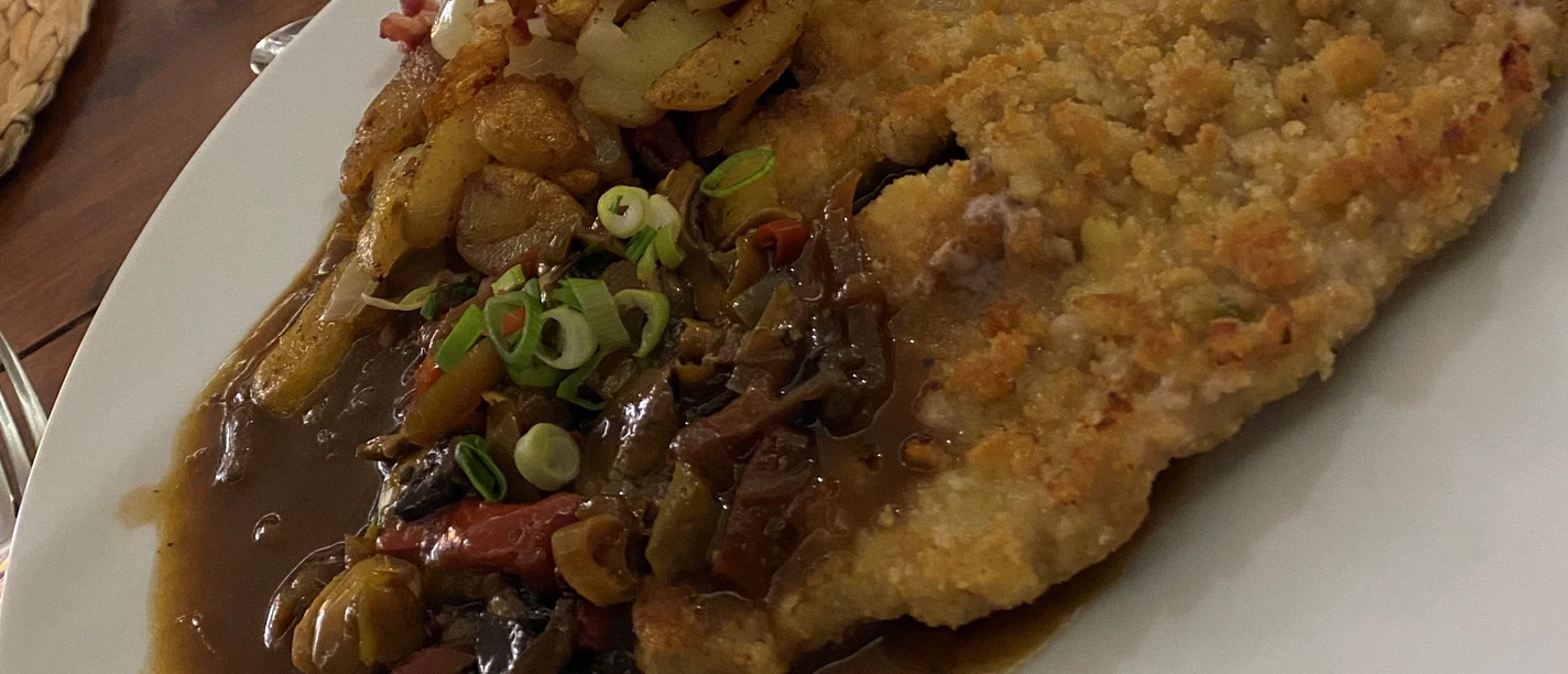 A plate with Jägerschnitzel, fried potatoes, gravy and vegetable garnish, next to it a glass of red wine.
