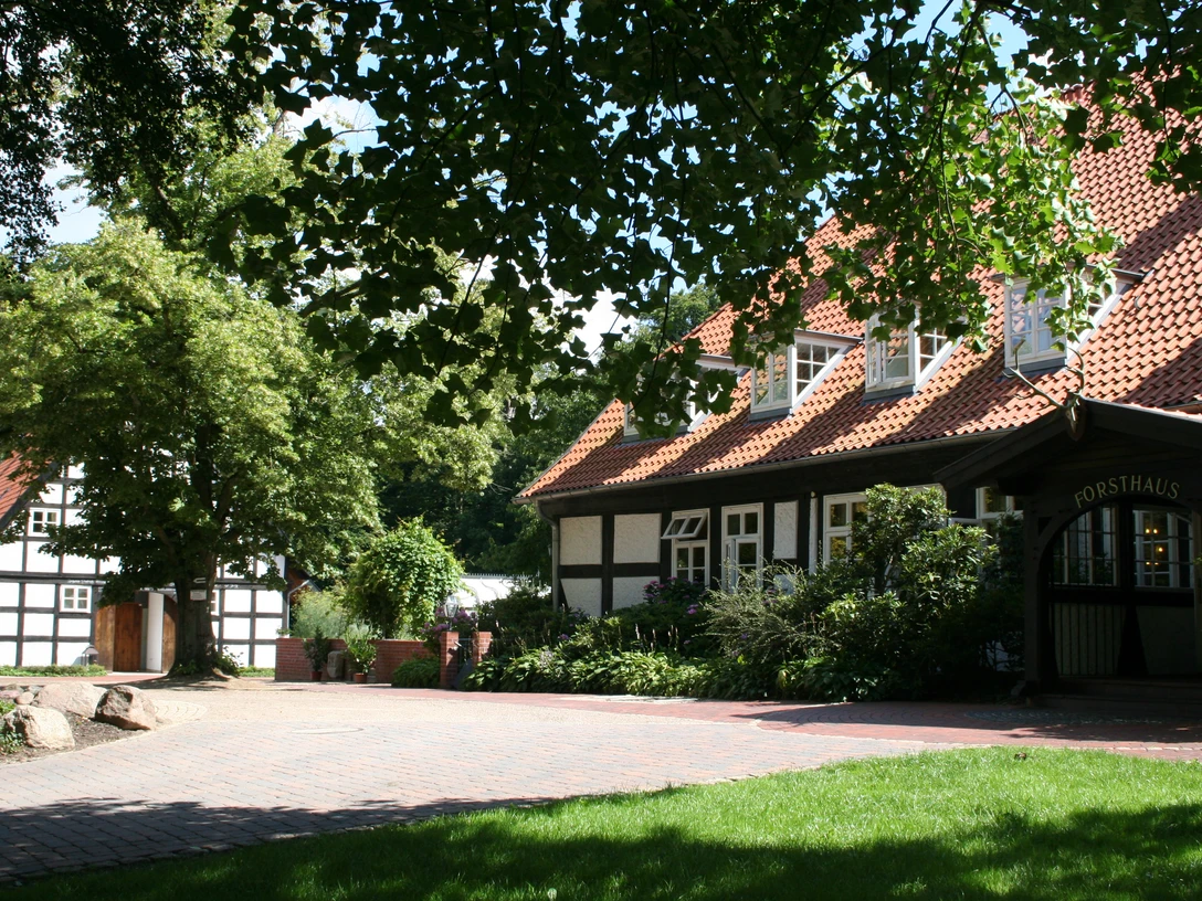 Bruchhausen-Vilsen Forsthaus Heiligenberg (12).JPG Ein traditionelles Fachwerkgebäude mit roten Dächern, umgeben von grünen Bäumen und Rasenflächen.