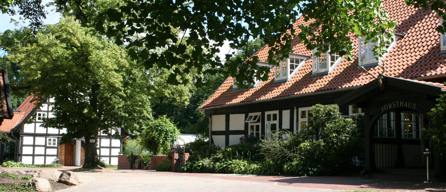 Bruchhausen-Vilsen Forsthaus Heiligenberg (12).JPG Ein traditionelles Fachwerkgebäude mit roten Dächern, umgeben von grünen Bäumen und Rasenflächen.