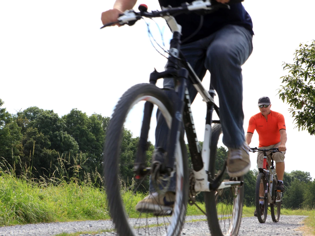 Ausflug auf dem PanoramaRadweg niederbergbahn Zwei Personen radeln entlang eines Schotterwegs, umgeben von grüner Vegetation, bei sonnigem Wetter.