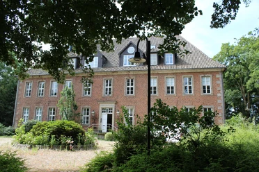 Ein historisches Backsteingebäude mit Sprossenfenstern und Garten, von Bäumen und Grün umgeben.