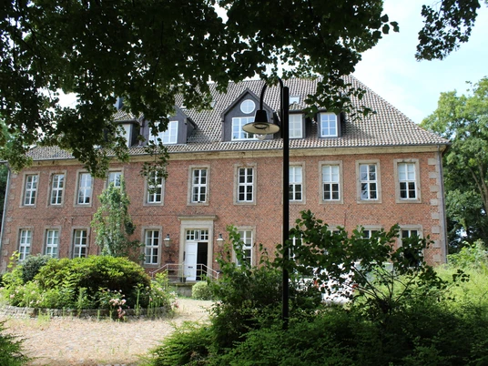 Ein historisches Backsteingebäude mit Sprossenfenstern und Garten, von Bäumen und Grün umgeben.