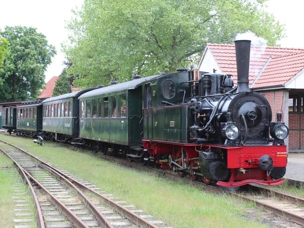 Museumseisenbahn Bruchhausen-Vilsen Eine historische Dampflok zieht grüne Waggons entlang eines Gleises durch eine grüne, ländliche Umgebung.