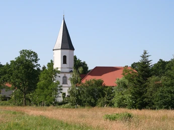 Eine weiße Kirche mit spitzem Turm schmiegt sich in eine grüne Landschaft und unter blauen Himmel.
