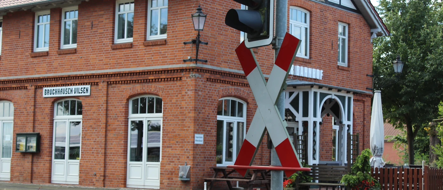 Ein rotes Backsteingebäude des Bahnhofs Bruchhausen-Vilsen mit charakteristischen weißen Fensterrahmen und einem Bahnübergangsschild im Vordergrund.