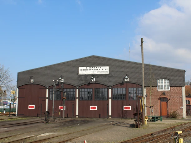 Museumseisenbahn-Depot mit historischen Schienenfahrzeugen vor strahlend blauem Himmel und Gleisanlagen.