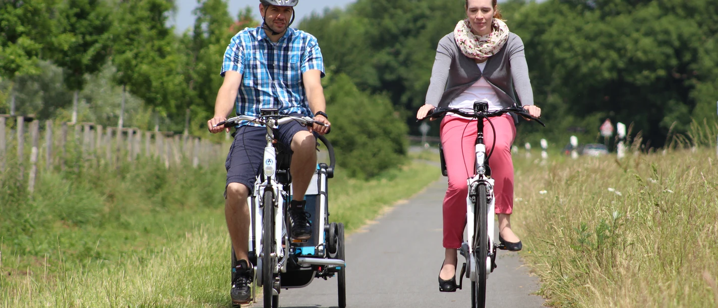 Zwei Radfahrer genießen eine Fahrt auf einem asphaltierten Weg, umgeben von grüner Natur und Bäumen.