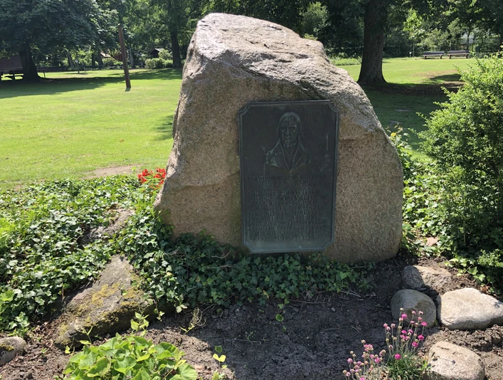 Gedenkstein aus Naturstein mit Gedenktafel in einem gepflegten Park, umgeben von blühenden Pflanzen.