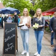 Zwei junge Frauen präsentieren frischen grünen Spargel auf einem belebten Markt unter Sonnenschirmen.