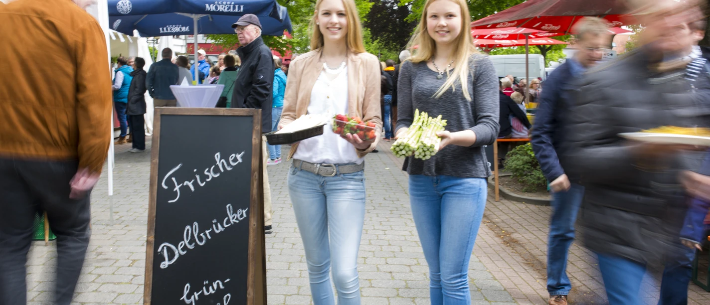 Zwei junge Frauen präsentieren frischen grünen Spargel auf einem belebten Markt unter Sonnenschirmen.