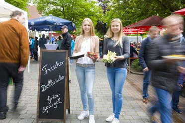 LOOS1855.jpg Zwei junge Frauen präsentieren frischen grünen Spargel auf einem belebten Markt unter Sonnenschirmen.