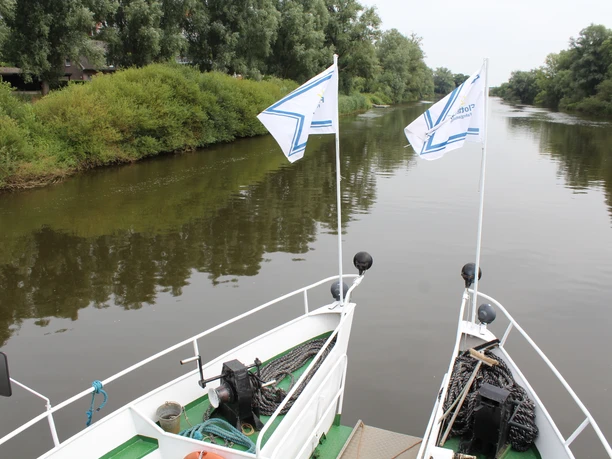 Zwei weiße Fahnen wehen über dem Bug eines Ausflugsschiffes auf einem ruhigen Fluss.