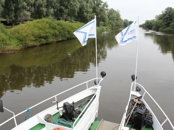 MS "Stadt Verden" Zwei weiße Fahnen wehen über dem Bug eines Ausflugsschiffes auf einem ruhigen Fluss.