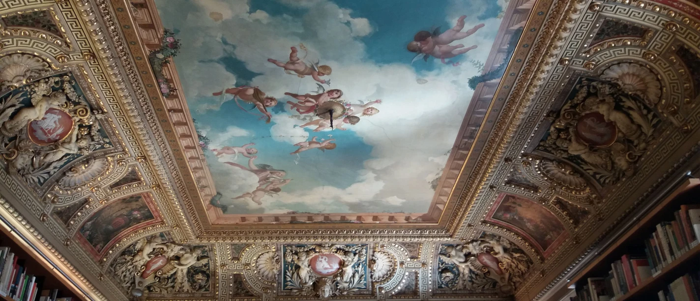 Bibliothek mit kunstvoller Decke aus einem Gemälde mit Engeln und Wolken und verschiedenen Ornamenten.