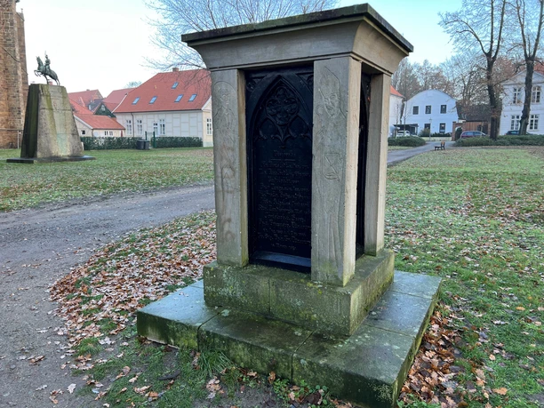 Bischofsdenkmal Verden Steinernes Denkmal mit filigran gearbeiteter Inschrift, umgeben von herbstlichem Laub im Park.