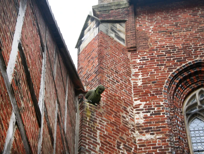 Ein Wasserspeier aus Stein ragt aus einer roten Backsteinwand unter einem gotischen Fenster hervor.