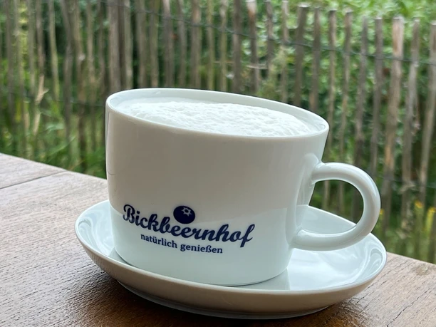 Witte mok met het Bickbeernhof-logo, gevuld met schuim, op een houten tafel tegen een groene achtergrond.