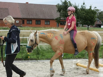 Reiten auf dem Herzfeldhof Ein Kind mit Helm reitet auf einem bunt bemalten Pferd auf einem Reitplatz, begleitet von einer Frau.