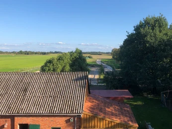 Über dem Herzfeldhof Ein historischer Hof mit Ziegelgebäude, bewaldetem Garten und angrenzendem Feld bei klarem Wetter.