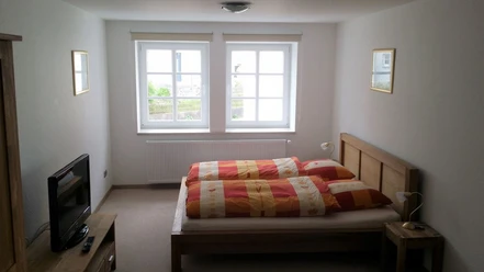 Kukuk4_web.jpg Helles Schlafzimmer mit zwei Fenstern, Doppelbett mit orange-rotem Bettzeug und Fernseher links.