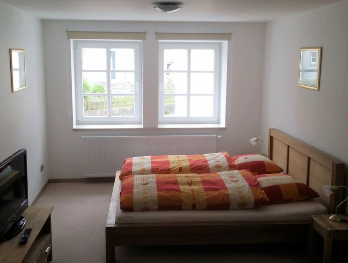 Kukuk4_web.jpg Helles Schlafzimmer mit zwei Fenstern, Doppelbett mit orange-rotem Bettzeug und Fernseher links.