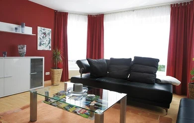 Nieheim-web-FW-Moeller-Merlsheim-02.jpg Ein modernes Wohnzimmer mit roter Wand, Ledersofa, Glastisch und großen Fenstern mit Tageslicht.