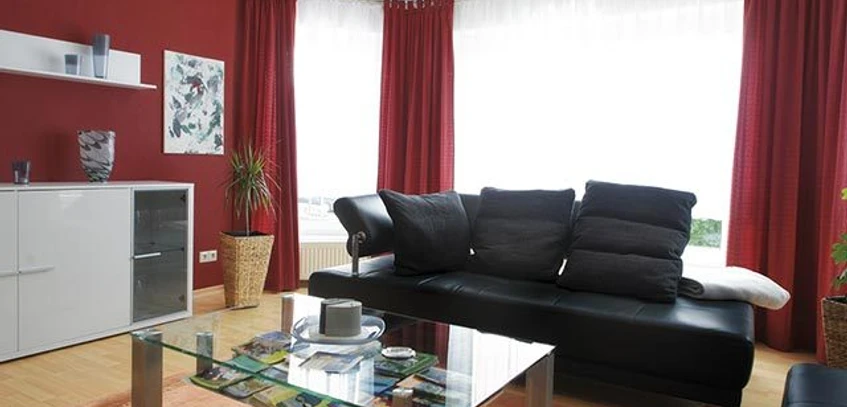 Nieheim-web-FW-Moeller-Merlsheim-02.jpg Ein modernes Wohnzimmer mit roter Wand, Ledersofa, Glastisch und großen Fenstern mit Tageslicht.