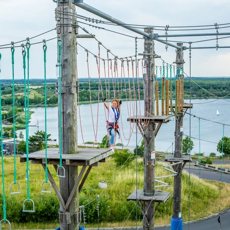 A woman climbs high above the ground in the climbing park at Lake Markkleeberg with a view of the lake, leisure activity, family outing