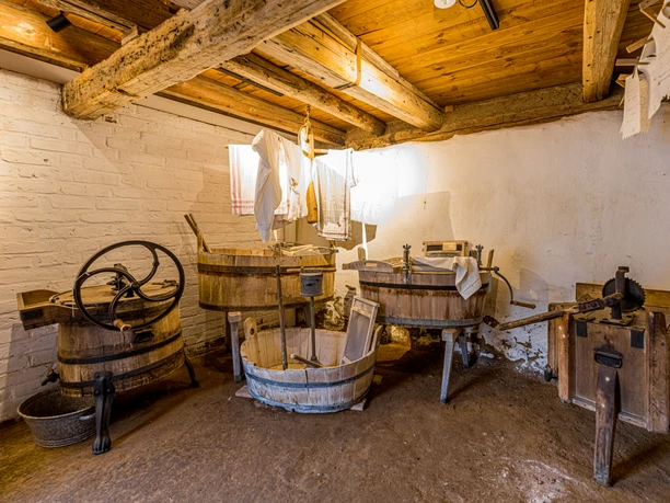 Geschichtenhof Wyhra Waschküche mit traditionellen Holzmollen, Wäscheleinen und rustikalem Holzgebälk an der Decke.