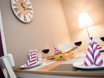 ferienwohnung_happiness-9_3000px.jpg Essbereich mit modernem, hell beleuchtetem Tisch, gedeckt mit Weingläsern, Käse, Trauben und Broten.