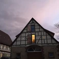 Ferienwohnung Historische Scheune - Außenansicht Fachwerkhaus im Abendlicht mit erleuchtetem Fenster und Wolken am Himmel, umgeben von kahlen Bäumen.