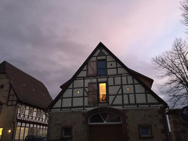 Ferienwohnung Historische Scheune - Außenansicht Fachwerkhaus im Abendlicht mit erleuchtetem Fenster und Wolken am Himmel, umgeben von kahlen Bäumen.