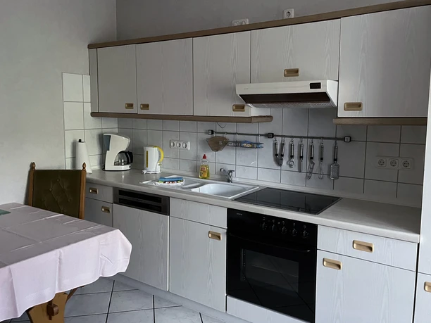 Apartment 03 Wetendorf Grau-weiße Küche mit Einbaugeräten, Doppelspüle und Esstisch mit weißem Tuch, Holzstühlen anbei.