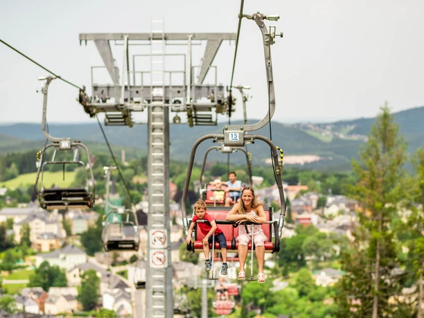 Wurzelrudi Sessellift Zwei Personen fahren im Wurzelrudi Sessellift über eine bewaldete Landschaft.