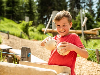 Wurzelrudi Murmelbahn Ein Junge mit roten Shirt hält lächelnd zwei Kugeln an einer Outdoor-Murmelbahn.