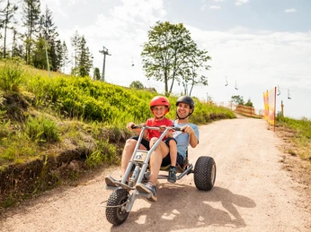 Wurzelrudi Mountaincart Vater und Sohn fahren mit einem dreirädrigen Mountaincart auf einem Schotterweg bergab.Father and son ride a three-wheeled mountain cart downhill on a gravel path.Otec a syn jedou na tříkolovém horském vozíku z kopce po štěrkové cestě.Ojciec i syn jadą trójkołowym wózkiem górskim w dół po żwirowym torze.Vader en zoon rijden met een driewielig bergkarretje bergafwaarts over een grindpad.Padre e figlio guidano un carrello da montagna a tre ruote in discesa su una pista di ghiaia.