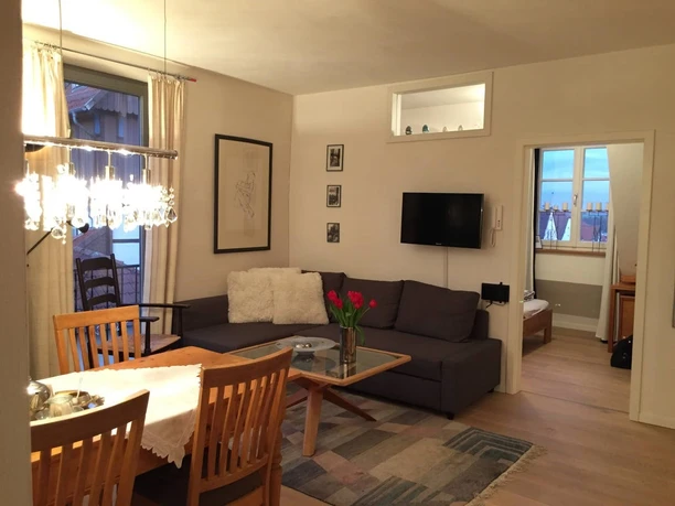 Ferienwohnung Historische Scheune_Wohnzimmer.jpg Wohnzimmer einer Ferienwohnung, mit grauem Sofa, Holztisch, Essbereich und dekorativen Lampen.