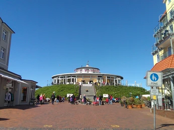 Eilanddorp Wangerooge Cafe Pudding.jpg Rundes Gebäude des Café Pudding auf einer Anhöhe mit Treppe, umgeben von Besuchern bei Sonnenschein.Round building of Café Pudding on a hill with stairs, surrounded by visitors in the sunshine.Edificio rotondo del Café Pudding su una collina con scale, circondato da visitatori sotto il sole.Rond gebouw van Café Pudding op een heuvel met trappen, omringd door bezoekers in de zon.Okrągły budynek Café Pudding na wzgórzu ze schodami, otoczony gośćmi w promieniach słońca.Clădire rotundă a Café Pudding pe un deal cu scări, înconjurată de vizitatori în soare.Café Puddings runde bygning på en bakke med trapper, omgivet af besøgende i solskin.