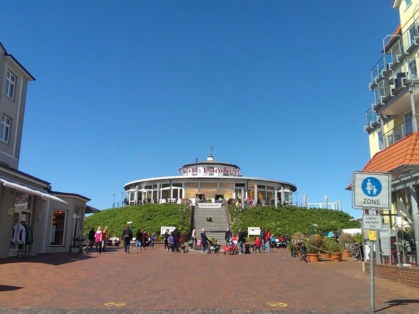 Eilanddorp Wangerooge Cafe Pudding.jpg Rond gebouw van Café Pudding op een heuvel met trappen, omringd door bezoekers in de zon.