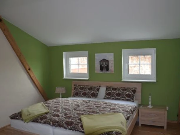 Ferienwohnung Böhlen - Schlafzimmer Modernes Schlafzimmer mit Doppelbett, grünen Wänden und Holzbalken. Zwei Fenster spenden Licht.
