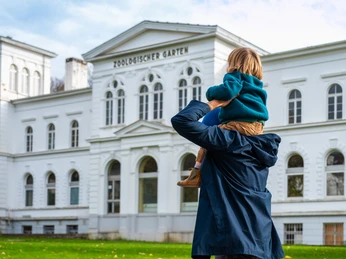 Der Grüne Zoo Wuppertal Eine Person mit Kind vor dem Gebäude des Zoologischen Gartens