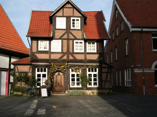 A traditional half-timbered house with the words "Weinhaus" on it, surrounded by brick buildings, under a blue sky.