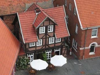 Fachwerkhaus mit rotem Ziegeldach, flankiert von roten Backsteingebäuden, davor Sonnenschirme.